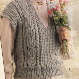 Puede incluir: Un chaleco de punto gris sin mangas con cable y cuello en V. El chaleco se lleva sobre una blusa blanca con cuello de volantes.