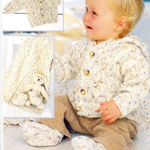 Puede incluir: Un bebé con un cárdigan de punto color crema con botones grandes de madera, botines a juego y una manta. El conjunto tiene un patrón moteado con acentos marrones y amarillos, y se usa con pantalones de pana marrones.