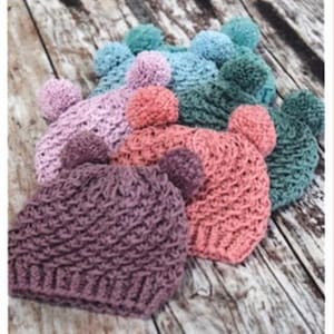 May include: A collection of handmade crochet beanies in various colors, including purple, pink, coral, teal, and blue. Each beanie features two ear-shaped pom-poms on top. The hats are arranged on a wooden surface.
