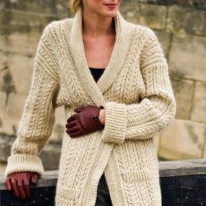 Może przedstawiać: Kremowy, długi kardigan z dzianiny z kołnierzem szalowym i kieszeniami. Kardigan ma wzorzysty wzór dzianiny i jest noszony z ciemnoczerwonymi rękawiczkami. Rękawy mają prążkowane mankiety. Ubranie jest noszone na czarnym topie i ciemnych spodniach.