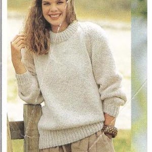 May include: A light beige knit sweater with a crew neck and long sleeves. The sweater is paired with tan-colored pleated pants. The image has a soft, natural lighting and a vintage aesthetic.