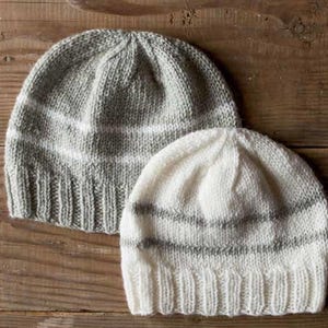 May include: Two knitted beanies in the image. One is gray with white stripes, the other is white with gray stripes. The hats are made of a soft, textured yarn and are likely handmade. The beanies are displayed on a wooden surface.