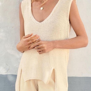 Può includere: Top senza maniche in maglia color crema con scollo a V profondo e spacchi laterali. Il top è realizzato in un tessuto a maglia strutturato ed è abbinato a pantaloni color crema. È visibile una collana d'oro.