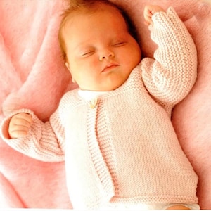May include: A sleeping baby wearing a pale pink knitted cardigan with a single button. The baby lies on a soft, pink blanket. The cardigan features ribbed cuffs and a textured knit pattern.