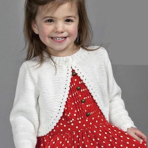 May include: A young child wearing a white, knitted bolero sweater over a red polka dot dress. The sweater has a scalloped edge and button closure. The dress features a round neckline and green buttons.