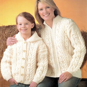 Op de afbeelding: Twee mensen dragen witte gebreide truien met kabelbreipatronen. De trui aan de linkerkant heeft een capuchon en knopen. De trui aan de rechterkant heeft een rits. Beide truien zijn gemaakt met een dik garen.