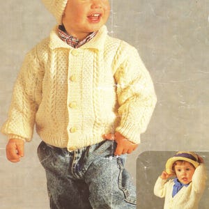 May include: A young child wearing a white knitted cardigan with a cable knit pattern and a white knitted hat. The child is wearing blue jeans and is standing on a light gray background.