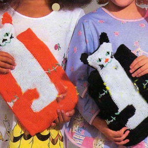 May include: Two knitted cat-shaped pillows. One is orange and white, the other is black and white. The pillows have embroidered facial features and are held by children. The image has a vintage, handmade aesthetic.