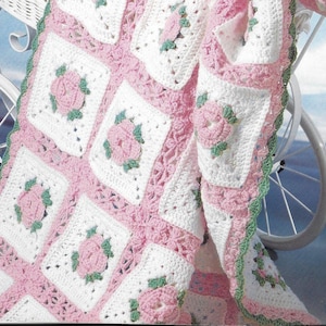 Op de afbeelding: Een handgemaakte gehaakte deken met een herhalend patroon van witte vierkanten, elk met een roze roos en groene bladeren. De vierkanten zijn verbonden door roze haakwerk en de deken heeft een groene geschulpte rand. De deken is over een witte kinderwagen gedrapeerd.