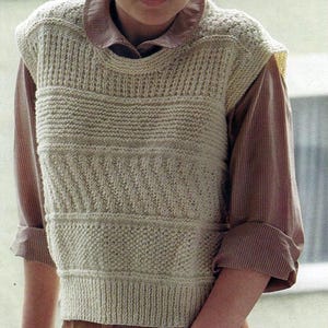 Puede incluir: Un chaleco de punto color crema con un patrón texturizado. El chaleco se lleva sobre una camisa con cuello con un patrón de rayas marrones y blancas. La persona lleva pantalones de pana marrones.