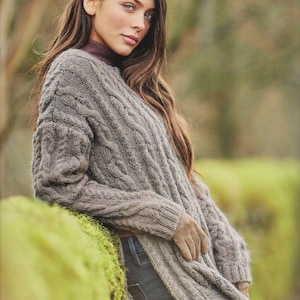 Könnte beinhalten: Eine Frau trägt einen grauen Strickpullover mit Zopfmuster und einem langen, fließenden Design. Der Pullover hat eine lockere Passform und wird mit einer blauen Jeans getragen. Die Frau steht vor einer grünen, moosbewachsenen Wand.