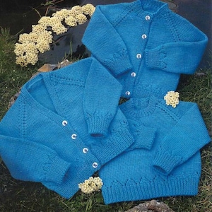 May include: Three hand-knitted, turquoise blue sweaters are displayed on a grassy surface. Two are cardigans with button closures, and one is a crew neck sweater. The sweaters have ribbed cuffs and hems, and are adorned with small, white flowers.
