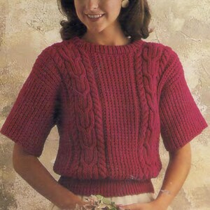 May include: A red cable knit jumper with short sleeves. The jumper has a round neckline and a ribbed hem.