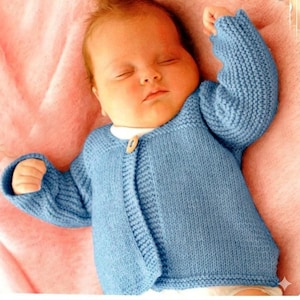 May include: A sleeping baby wearing a pale blue knitted cardigan with a single wooden button. The baby is on a pink blanket. The cardigan has ribbed cuffs and a round neckline. The cardigan is made of a soft, textured yarn.