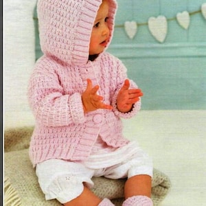 May include: A baby wearing a light pink knitted hooded jacket with ear details, matching booties, and a button-up closure. The baby is also wearing white shorts. The outfit is made of a soft, textured yarn. The baby is sitting on a blanket.