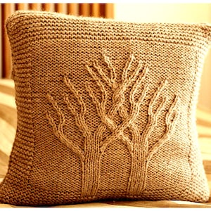 May include: A square, knitted pillow in a warm brown color. The pillow features a detailed tree design in the center, with the branches and trunk created using a textured knit stitch. The pillow is a cozy home decor item.