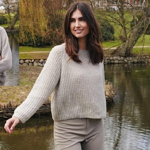 Può includere: Una donna indossa un maglione a coste grigio chiaro e pantaloni beige vicino a uno specchio d'acqua. Il maglione ha uno scollo tondo e maniche lunghe. Lo sfondo comprende alberi e un muro di pietra.