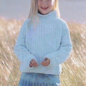 May include: A light blue turtleneck sweater with a textured knit design. The sweater is paired with a denim skirt. The person is smiling and standing in a field of tall grass.