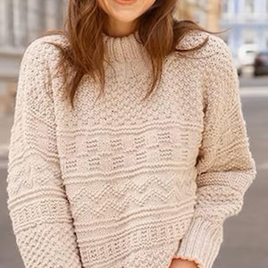 Puede incluir: Un suéter de punto beige con cuello alto y mangas largas. El suéter presenta un patrón geométrico en el pecho y las mangas. Los puños y el dobladillo acanalados añaden un toque clásico a esta prenda acogedora.