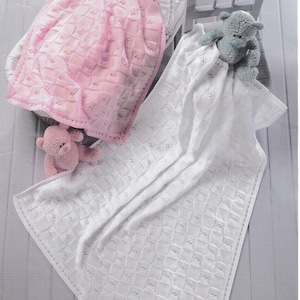 May include: A pink and white baby blanket set. The pink blanket is folded in a bassinet, and the white blanket is draped over a wooden crib. Both blankets have a diamond pattern. A pink and a gray teddy bear are also in the image.