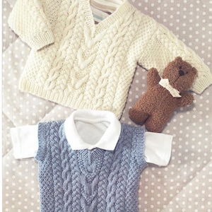 May include: Cream-colored cable-knit sweater and a blue cable-knit vest with a white collared shirt underneath. A small brown teddy bear with a white ribbon is also present. The items are arranged on a polka-dot patterned surface.