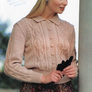 Puede incluir: Un cárdigan de punto beige claro con cuello y cierre de botones. El cárdigan presenta detalles de punto trenzado en las mangas y los paneles delanteros. El texto "40 Rolle Street Exmouth" es visible. La persona lleva una falda estampada.