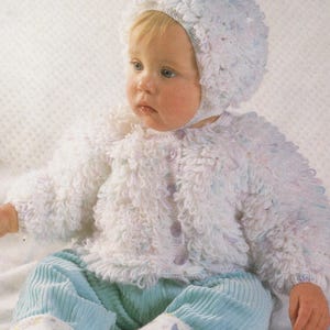 May include: A white crocheted baby jacket and hat set with a fluffy texture. The jacket has a button closure and the hat has a tie.