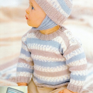 Op de afbeelding: Een baby draagt een gebreide trui en muts set. De trui en muts zijn gestreept in lichtblauw, wit en beige. De muts heeft een pompon en oorflappen. De trui heeft twee knopen op de schouder.