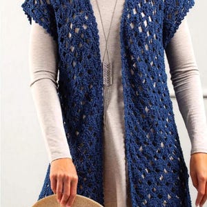 May include: A woman wearing a navy blue crochet vest over a long-sleeved gray shirt. The open-weave vest has a long, flowing design. She is holding a straw hat with a brown brim. A silver necklace hangs down.