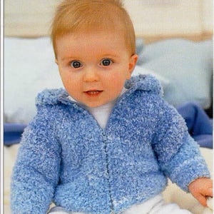 May include: A baby in a light blue, hooded, zip-up jacket. The jacket has a fluffy texture and is worn with white shorts. The baby has fair hair and is looking directly at the camera.