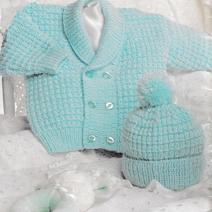 Puede incluir: Conjunto de bebé tejido en azul claro, que incluye una rebeca con cuello chal y cierre de botones, y un gorro a juego con pompón. El conjunto está hecho de un hilo suave y texturizado.