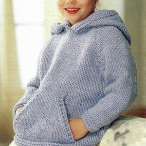 Peut inclure: Un sweat à capuche tricoté bleu clair avec une poche kangourou. Le pull à capuche est fait d'un fil épais et texturé. L'enfant porte le pull, les mains dans la poche avant.