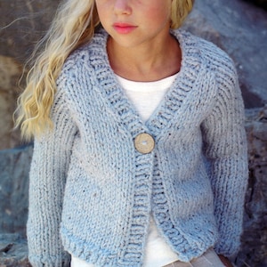 Peut inclure: Un cardigan tricoté bleu clair avec un col en V et un seul gros bouton en bois. Le pull a un motif de tricot texturé et des manches longues. Le cardigan est porté sur un haut blanc.