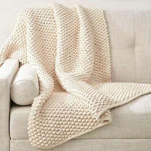 May include: A cream-coloured, chunky knit throw blanket draped over a light-coloured sofa. The blanket has a textured, woven appearance and is folded to display its thick, warm design. A cylindrical pillow rests on the sofa.