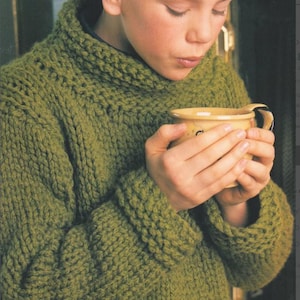 Könnte beinhalten: Eine Person trägt einen grob gestrickten olivgrünen Pullover mit hohem Kragen und gerippten Bündchen. Die Person hält eine gelbe Tasse mit gebogenem Henkel. Der Pullover scheint handgefertigt zu sein.