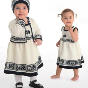 以下が含まれることがあります： ネイビーブルーのアクセントが付いた、お揃いのクリーム色の衣装を着た2人の幼い子供たち。一人は帽子、カーディガン、ドレス、靴を着用。もう一人はドレスを着用。衣装は雪の結晶のデザインが特徴です。