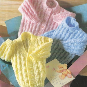 May include: Three hand-knitted baby sweaters in pastel colors: pink, blue, and yellow. The sweaters have a ribbed texture and button closures. A greeting card with a teddy bear illustration is also visible.