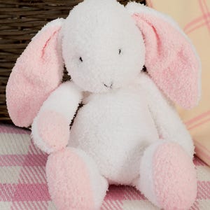 May include: A plush white bunny with pink inner ears and paw pads. The stuffed animal is sitting on a pink and white checkered surface. The bunny has a soft, textured appearance, and its features include small black eyes and a stitched nose.