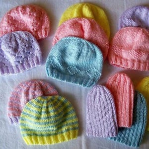 May include: A collection of colorful, knitted beanies in various shades of pink, yellow, blue, and purple. The hats are handmade, with different stitch patterns and textures. Some have striped designs, while others are solid colors.