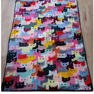 Op de afbeelding: Een handgemaakte gehaakte deken met een herhalend patroon van kattengezichten in verschillende kleuren. De deken heeft een zwarte rand en is gemaakt met garen in tinten roze, blauw, geel en groen.