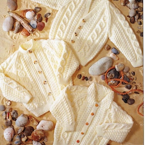 May include: Several cream-coloured cable-knit cardigans with button fastenings. The garments are displayed on a sandy surface with pebbles and seashells. The knitwear has textured patterns and is likely made of wool or a similar material.