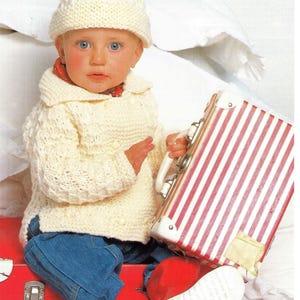 May include: A baby in a cream knitted hat and sweater, blue jeans, and red boots, holding a red and white striped suitcase. The outfit is suitable for cooler weather. The suitcase has a white handle.