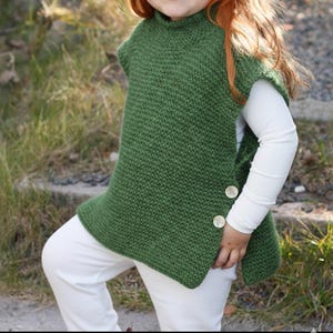 May include: A green knitted child's vest with a high collar and two large, round buttons. The vest is paired with white pants and a long-sleeved white shirt. The knitted texture is visible, and the vest appears to be handmade.