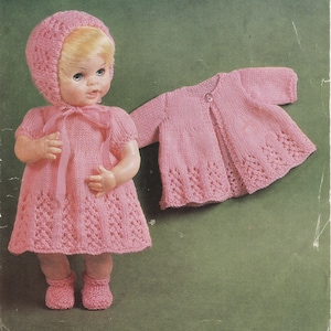 May include: A vintage doll wearing a knitted pink dress, bonnet, and booties. A matching pink cardigan lies beside the doll. The dress and cardigan feature a decorative openwork pattern. The doll has blonde hair and is posed against a green background.