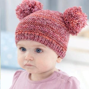 May include: A knitted baby hat in shades of pink and red, featuring two large pom-poms on top. The hat is made of a textured yarn and has a ribbed brim. The baby is wearing a pink long-sleeved shirt.