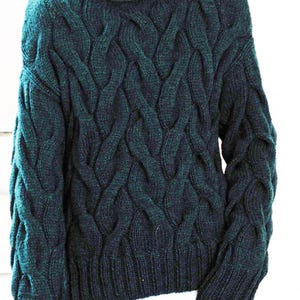 May include: Dark teal cable-knit sweater. The sweater features a thick, textured cable-knit pattern. The neckline is a crew neck, and the sleeves are long. The sweater is worn over a white collared shirt and blue jeans.