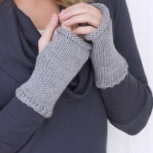 Könnte beinhalten: Ein Paar hellgraue gestrickte Fingerhandschuhe. Die Handschuhe haben eine gerippte Manschette und einen gewellten Rand. Die Handschuhe sind aus einem weichen, strukturierten Garn gefertigt. Die Handschuhe sollen die Hände warm halten, während die Finger frei bleiben.