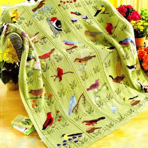 May include: A light green knitted blanket featuring various colorful bird designs. The blanket has a textured pattern with vertical lines and embroidered birds, flowers, and foliage. The blanket is draped over a chair, with a book titled "Birds" visible.