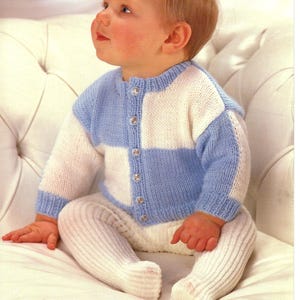 May include: A baby in a blue and white knitted cardigan with silver buttons, and white ribbed tights. The cardigan has a checkerboard pattern. The baby is seated on a white surface.