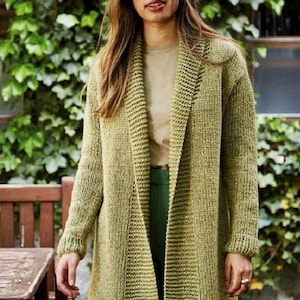 May include: A long, olive green knit cardigan with a shawl collar. The cardigan is worn over a light-colored top and green pants. The sleeves have ribbed cuffs. The background features lush green foliage and a wooden table.